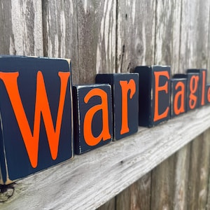 May include: Wooden blocks spelling "War Eagle" in orange letters on a navy blue background. The blocks are arranged on a wooden shelf against a weathered wood background. The letters are bold and easy to read.