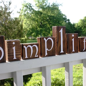 May include: Wooden blocks spelling out the word "Simplify" in white letters on a brown background. The blocks are arranged on a white railing.