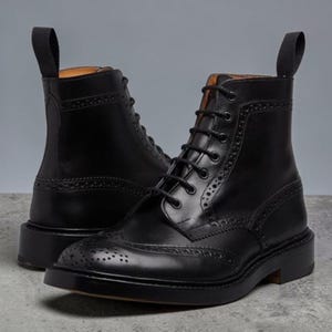 May include: A pair of black leather lace-up boots featuring brogue detailing, a wingtip toe cap, and a pull tab at the heel. The boots have a stacked heel, a rounded toe, and black laces threaded through metal eyelets, showcasing a polished finish.