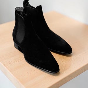 Puede incluir: Un par de botines Chelsea negros de ante con punta afilada y tacón bajo. Los botines se exhiben sobre una superficie de madera clara. Tienen paneles laterales elásticos y una lengüeta en la parte trasera.