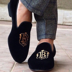 May include: Black velvet loafers with gold embroidered emblem. The shoes have a classic slip-on design and are paired with grey plaid trousers. The emblem features the letters "B" and "L" in an ornate script.