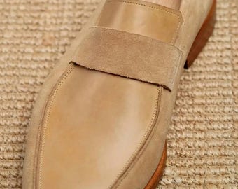 May include: A tan suede loafer featuring a smooth leather strap across the vamp, a square toe, and a brown stacked heel. The shoe includes a wooden shoe tree inserted inside to maintain its shape, showcasing a clean, structured design.