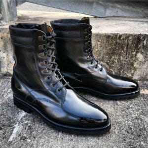 May include: A pair of glossy black leather boots with a lace-up design. The boots have a high-top style with a smooth, polished finish. The soles are black, and the boots appear to be new.