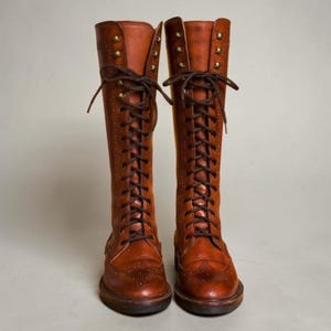 May include: A pair of knee-high tan leather boots featuring a full lace-up front with dark brown laces, brass-toned eyelets, and decorative brogue detailing along the toe cap and side panels, set on a low heel with a rounded toe silhouette.