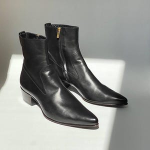 May include: A pair of black leather ankle boots with pointed toes and a low heel. The boots have a zipper closure on the side and a gold-colored zipper pull. The boots are displayed on a white surface.