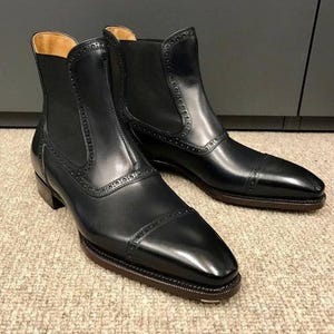 May include: A pair of black leather Chelsea boots with a polished finish. The boots have a classic design with a rounded toe, elastic side panels, and decorative stitching. The soles are dark brown.