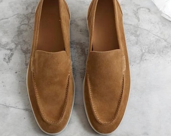Könnte beinhalten: Ein Paar hellbraune Wildleder-Slipper mit abgerundeter Zehenpartie, zum Hineinschlüpfen und mit sichtbaren Nähten am Vorderblatt. Die Schuhe haben ein glattes Innenfutter und eine dünne weiße Sohle, dargestellt aus der Vogelperspektive auf hellem Grund.