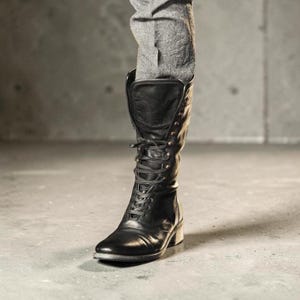 Puede incluir: Una bota de cuero negro a media pantorrilla con cierre frontal de cordones finos negros, punta redondeada, tacón bajo en bloque y una caña alta y estructurada que se extiende hacia arriba, usada con pantalones grises metidos en la bota.