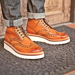 May include: A pair of tan leather brogue boots with white soles. The boots have decorative perforations and black laces. The boots are worn with blue denim jeans with the cuffs rolled up.