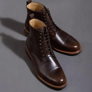 May include: A pair of dark brown leather boots with a cap toe design. The boots feature a lace-up closure and a light brown sole. The leather has a polished finish, set against a grey background.