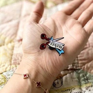 May include: A red and black scissors pin with a blue banner that reads "maker" is held in a hand. A gold bracelet with red clover charms is on the wrist. A floral mug and a quilted placemat are in the background.
