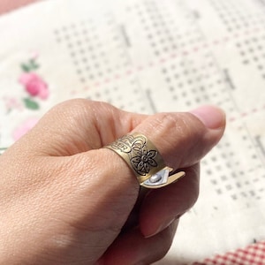 May include: A brass-colored thimble ring with a floral design and a small blade. The ring is worn on a finger, with a white and red checkered fabric in the background. The thimble ring is a sewing tool.