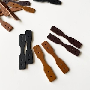 May include: An assortment of leather tags in various shades of brown and black, scattered on a white surface. The tags have a paddle-like shape with two holes at each end, suitable for crafting or labeling.