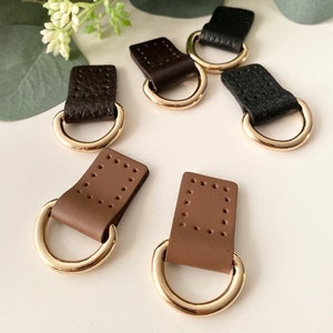 May include: A collection of leather and metal D-ring connectors in various shades of brown and black. Each connector features a rectangular leather tab with a series of small holes and a gold-colored metal D-ring. The connectors are designed for crafting and accessories.