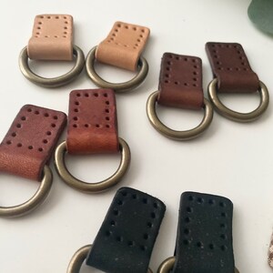 Puede incluir: Una colecci&oacute;n de anillas en D de cuero en varios colores, incluyendo beige, marr&oacute;n y negro. Cada anilla en D tiene un anillo met&aacute;lico unido a una pieza rectangular de cuero con detalles de costura. Dispuestas sobre una superficie blanca.