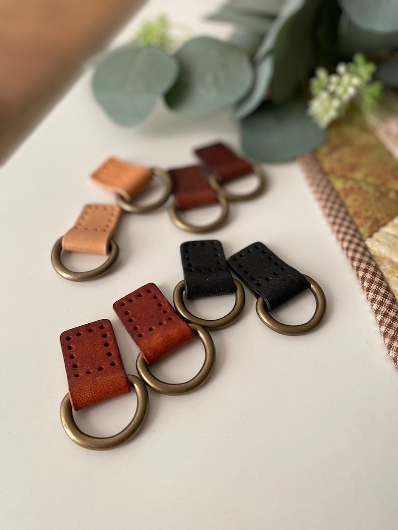 Puede incluir: Una colecci&oacute;n de presillas de cuero en varios colores, incluyendo beige, marr&oacute;n y negro, cada una unida a un anillo de metal color lat&oacute;n. Las piezas de cuero tienen detalles cosidos y los anillos son circulares.