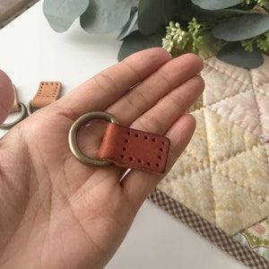 Puede incluir: Primer plano de un llavero de cuero con un anillo de lat&oacute;n. El llavero es de color marr&oacute;n rojizo y tiene forma rectangular con costuras decorativas. El anillo de lat&oacute;n est&aacute; unido a la parte superior de la pieza de cuero.
