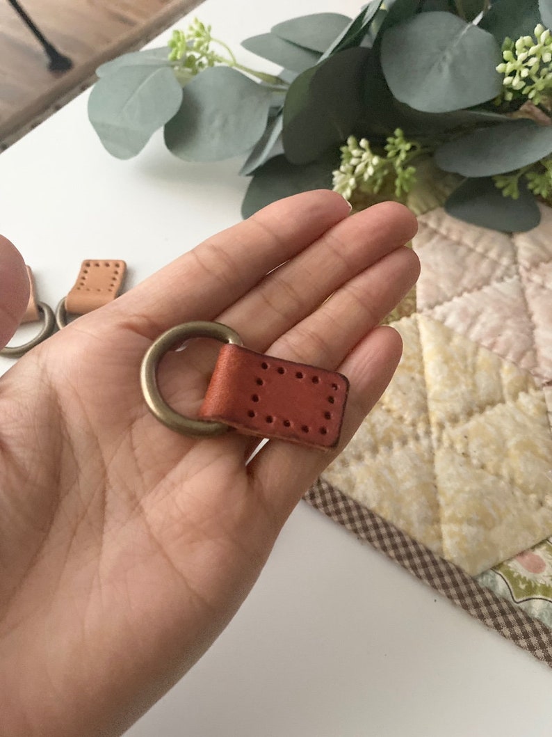 Puede incluir: Primer plano de un llavero de cuero con un anillo de lat&oacute;n. El llavero es de color marr&oacute;n rojizo y tiene forma rectangular con costuras decorativas. El anillo de lat&oacute;n est&aacute; unido a la parte superior de la pieza de cuero.