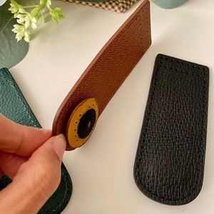 May include: Close-up of several leather magnetic snap closures in various colors. A brown closure with a yellow and black circular accent is held up, showcasing the design. Other closures are green and black, with a textured finish. These are ideal for handmade bags and accessories.