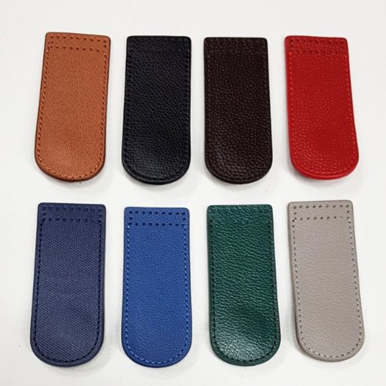May include: An assortment of eight leather magnetic bookmarks in various colors. The bookmarks are rectangular with rounded bottoms and stitched edges. Colors include brown, black, dark brown, red, navy, blue, green, and gray.