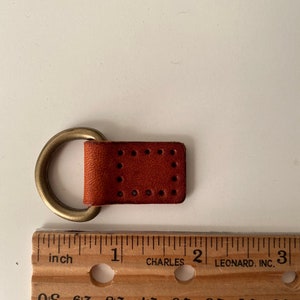 Puede incluir: Un peque&ntilde;o trozo de cuero marr&oacute;n rojizo rectangular con un anillo de lat&oacute;n adjunto. El cuero tiene una serie de agujeros espaciados uniformemente. Una regla de madera con marcas en pulgadas est&aacute; al lado.