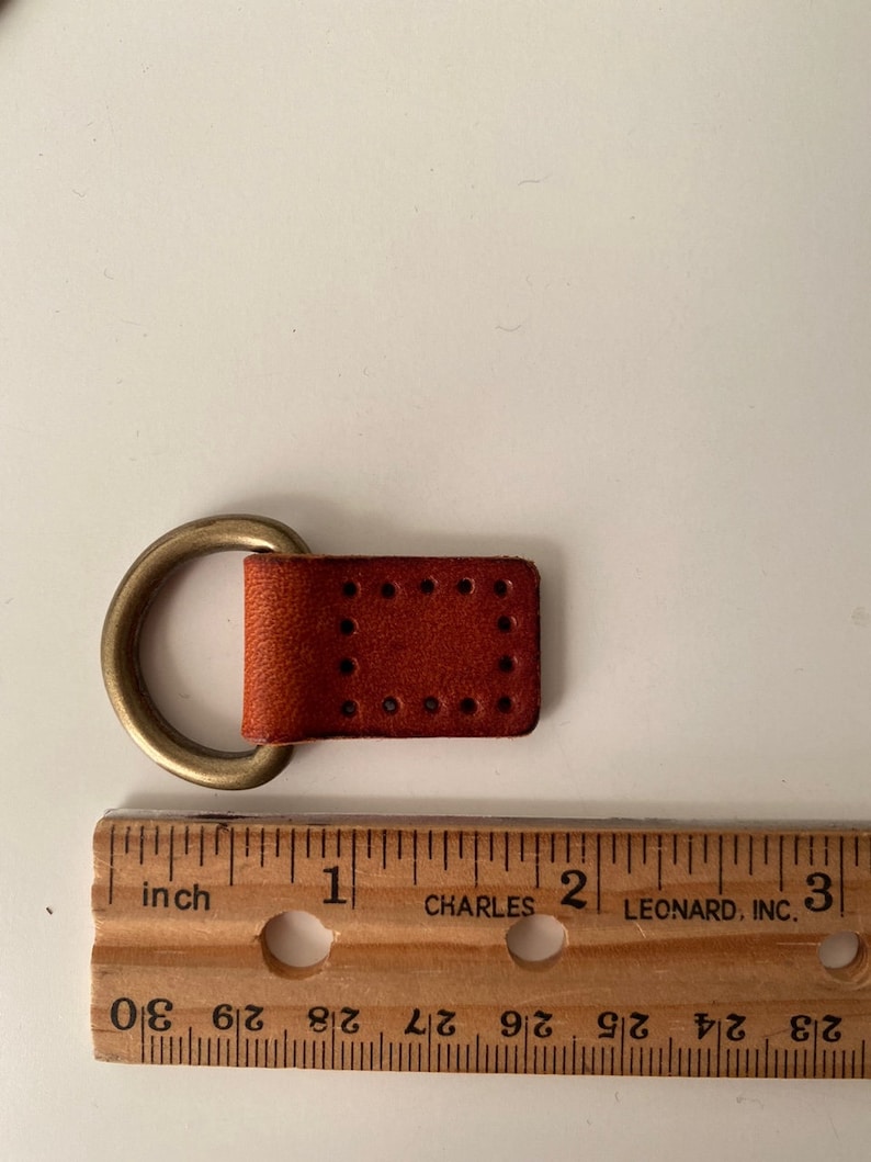 Puede incluir: Un peque&ntilde;o trozo de cuero marr&oacute;n rojizo rectangular con un anillo de lat&oacute;n adjunto. El cuero tiene una serie de agujeros espaciados uniformemente. Una regla de madera con marcas en pulgadas est&aacute; al lado.