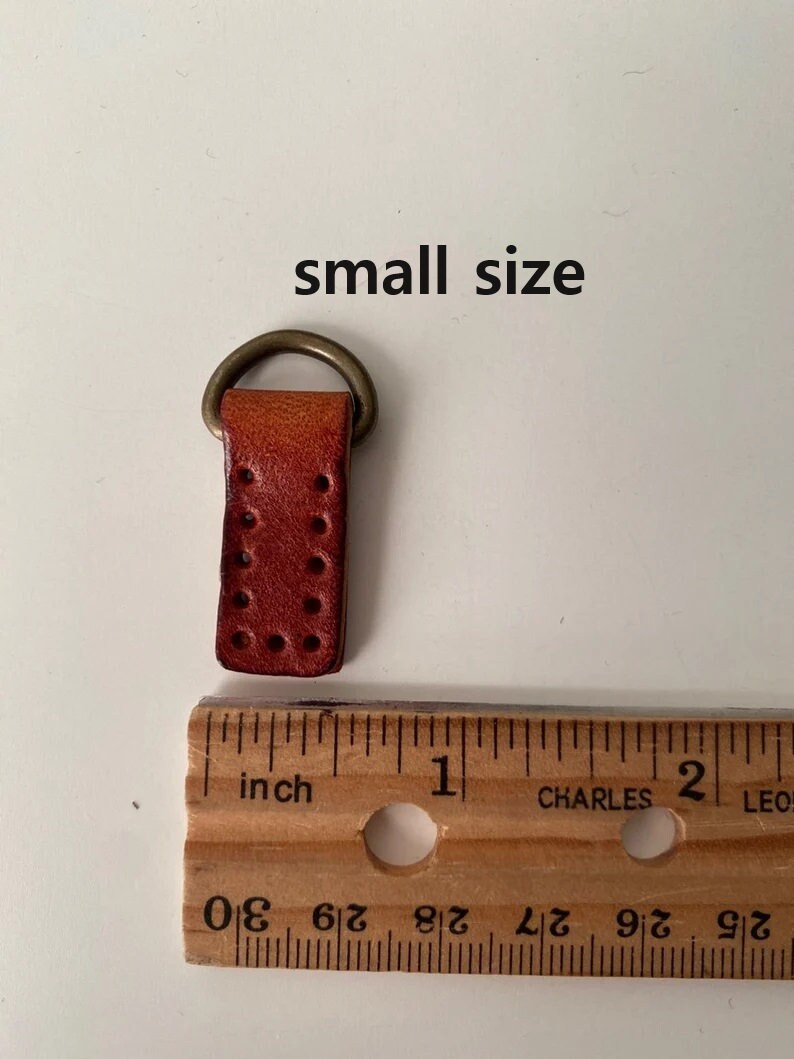 Puede incluir: Peque&ntilde;a correa de cuero marr&oacute;n rojizo con un anillo de metal color lat&oacute;n. La correa tiene varios agujeros peque&ntilde;os y espaciados uniformemente. El texto "small size" est&aacute; arriba. Una regla muestra unos 2,5 cm de largo.