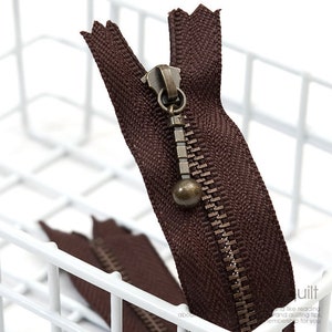 May include: Close-up of a brown zipper with a bronze-colored pull. The zipper teeth are a matching bronze color. The zipper is partially open, revealing the teeth and the fabric backing. The zipper is likely for sewing or crafting projects.