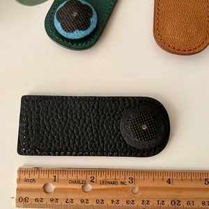 May include: A black leather needle minder with a textured surface and a round, black button. A green leather needle minder with a blue and black floral button is also visible. A ruler shows the needle minder is approximately 3 inches long.