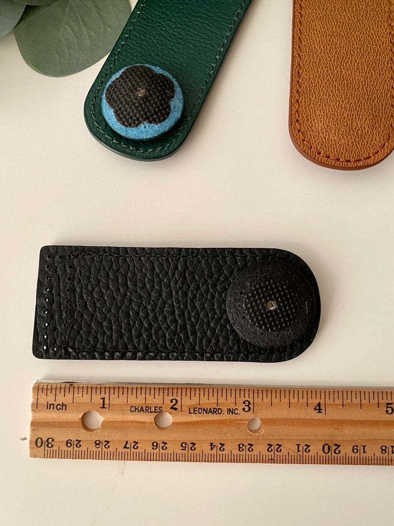 May include: A black leather needle minder with a textured surface and a round, black button. A green leather needle minder with a blue and black floral button is also visible. A ruler shows the needle minder is approximately 3 inches long.