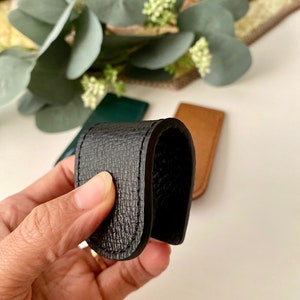 May include: A black leather money clip with a textured surface. The clip is held in a hand, with other leather items in green and brown visible in the background. The clip is designed to hold folded bills.