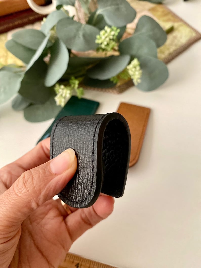 May include: A black leather money clip with a textured surface. The clip is held in a hand, with other leather items in green and brown visible in the background. The clip is designed to hold folded bills.