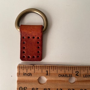 Puede incluir: Un peque&ntilde;o lazo de cuero marr&oacute;n con un anillo de metal color lat&oacute;n adjunto. La pieza de cuero tiene agujeros espaciados uniformemente y mide aproximadamente 2,5 cm de largo. Una regla de madera est&aacute; al lado del lazo de cuero.
