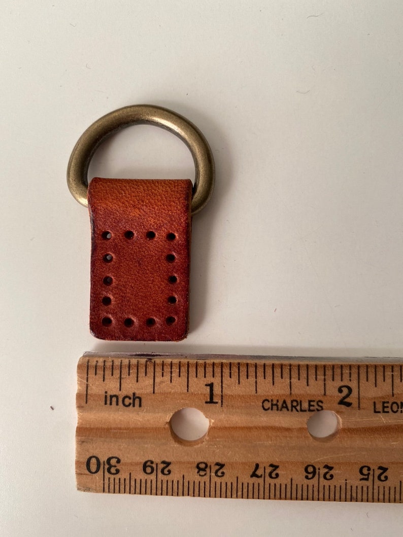 Puede incluir: Un peque&ntilde;o lazo de cuero marr&oacute;n con un anillo de metal color lat&oacute;n adjunto. La pieza de cuero tiene agujeros espaciados uniformemente y mide aproximadamente 2,5 cm de largo. Una regla de madera est&aacute; al lado del lazo de cuero.