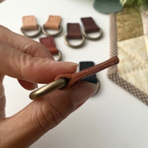 Puede incluir: Primer plano de varias leng&uuml;etas de cuero con anillos met&aacute;licos de color lat&oacute;n. Las leng&uuml;etas son de varios tonos de marr&oacute;n y beige. Una leng&uuml;eta se sostiene en la mano, mostrando el detalle del cuero y el anillo. El fondo est&aacute; borroso.