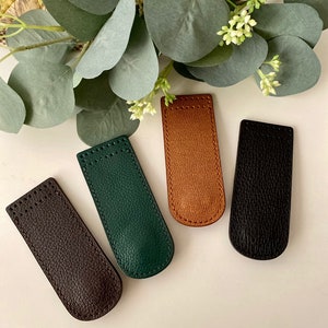 May include: Four leather pen sleeves in brown, green, tan, and black. Each sleeve is rectangular with a curved top and a textured surface. The sleeves are arranged on a white surface with green foliage in the background.