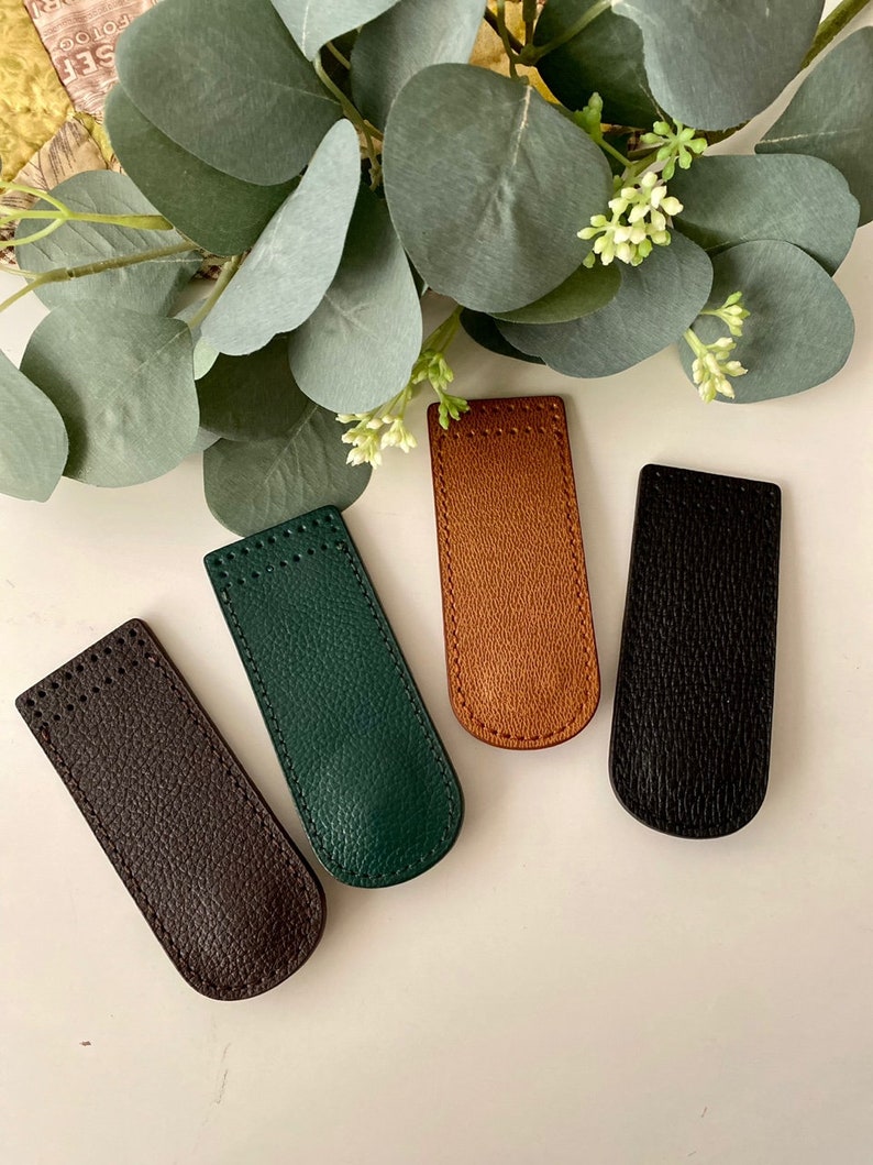 May include: Four leather pen sleeves in brown, green, tan, and black. Each sleeve is rectangular with a curved top and a textured surface. The sleeves are arranged on a white surface with green foliage in the background.