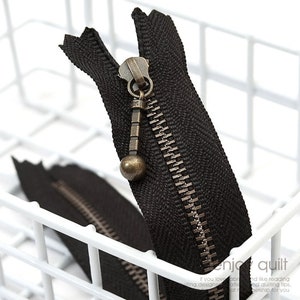 May include: Close-up of a black zipper with a bronze-colored pull tab and teeth. The zipper is partially open, revealing the teeth and the fabric backing. The zipper is in a white wire basket.