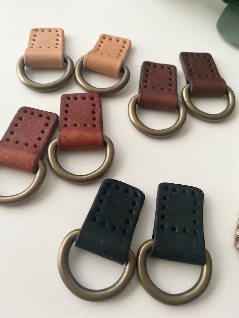 Puede incluir: Una colecci&oacute;n de anillas en D de cuero en varios colores, incluyendo beige, marr&oacute;n y negro. Cada anilla en D tiene un anillo met&aacute;lico unido a una pieza rectangular de cuero con detalles de costura. Dispuestas sobre una superficie blanca.