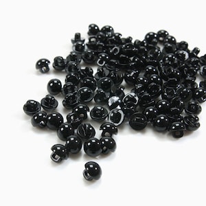 May include: A pile of small, shiny black buttons with a mushroom-shaped design. These buttons are made of a glossy material and have a loop on the back for sewing. They are scattered on a white surface.