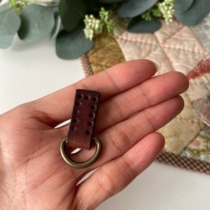 Puede incluir: Una peque&ntilde;a correa de cuero marr&oacute;n oscuro con un anillo de metal color lat&oacute;n. El cuero tiene varios agujeros peque&ntilde;os y espaciados uniformemente. El anillo est&aacute; unido a la parte inferior de la correa. El art&iacute;culo se sostiene en una mano.