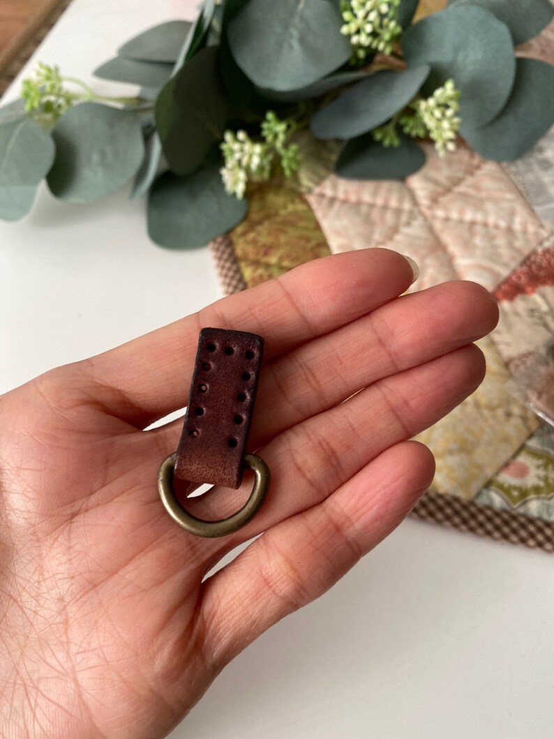 Puede incluir: Una peque&ntilde;a correa de cuero marr&oacute;n oscuro con un anillo de metal color lat&oacute;n. El cuero tiene varios agujeros peque&ntilde;os y espaciados uniformemente. El anillo est&aacute; unido a la parte inferior de la correa. El art&iacute;culo se sostiene en una mano.