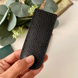 May include: A black leather rectangle with a textured surface is held in a hand. The leather has a subtle, patterned grain. The item is rectangular with rounded edges. The background includes greenery and a white surface.