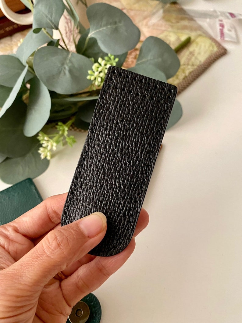 May include: A black leather rectangle with a textured surface is held in a hand. The leather has a subtle, patterned grain. The item is rectangular with rounded edges. The background includes greenery and a white surface.