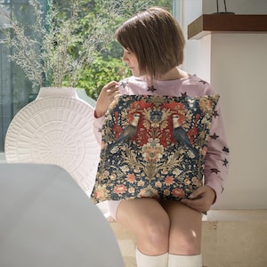 Puede incluir: Una persona joven sentada en una silla blanca, sosteniendo una almohada decorativa con un patrón floral y de pájaros. La almohada tiene un fondo azul oscuro con acentos rojos, dorados y verdes. La persona lleva una camisa rosa con estrellas blancas.