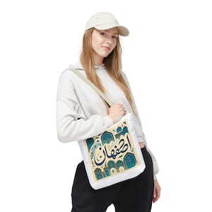 Puede incluir: Bolso tote blanco con un diseño turquesa y dorado con caligrafía árabe. El bolso tiene una correa beige para el hombro. La persona lleva una sudadera con capucha color crema y una gorra de béisbol.
