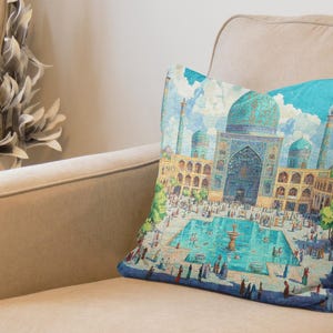Puede incluir: Un cojín decorativo con una ilustración detallada de un complejo arquitectónico de Oriente Medio con cúpulas turquesas y una piscina central. El cojín tiene un borde azul y está colocado sobre un sofá beige.