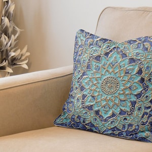 Puede incluir: Una almohada decorativa con un patrón de mandala azul y dorado. La almohada está hecha de una tela suave y texturizada y presenta detalles intrincados.