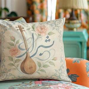 May include: Square decorative pillow with a watercolor design. The pillow features a tan stringed instrument, pink roses, and blue script on a cream background. The pillow is on a floral patterned chair.