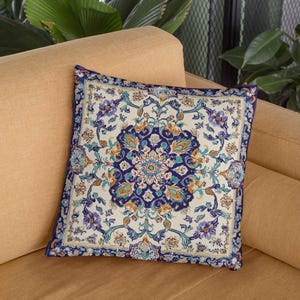 Puede incluir: Una almohada decorativa con un patrón floral azul, blanco y dorado. La almohada es cuadrada y tiene un fondo blanco con un borde azul.